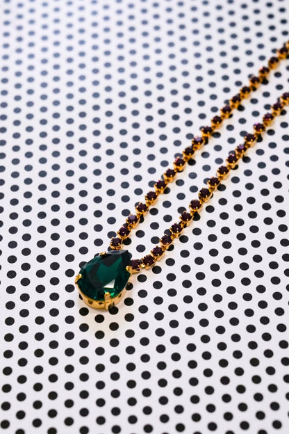 A gold necklace with a large green marquise-cut gemstone pendant and smaller gold and dark brown beads along the chain, displayed on a white background with a black polka dot pattern.