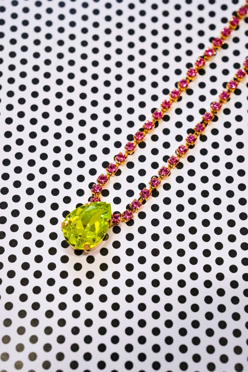 A gold necklace with a faceted lime green pendant and a chain accented with small pink gemstones, displayed on a white background with a black polka dot pattern.