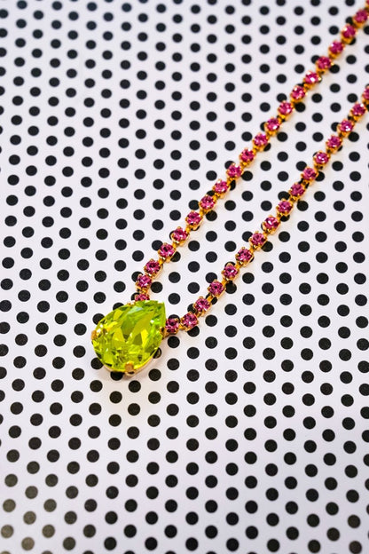 A gold necklace with a faceted lime green pendant and a chain accented with small pink gemstones, displayed on a white background with a black polka dot pattern.
