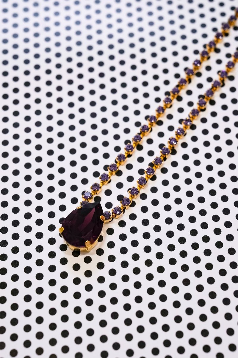 A gold chain necklace with a large teardrop-shaped dark purple gemstone pendant, accented with small purple stones, displayed on a white background with a black polka dot pattern.