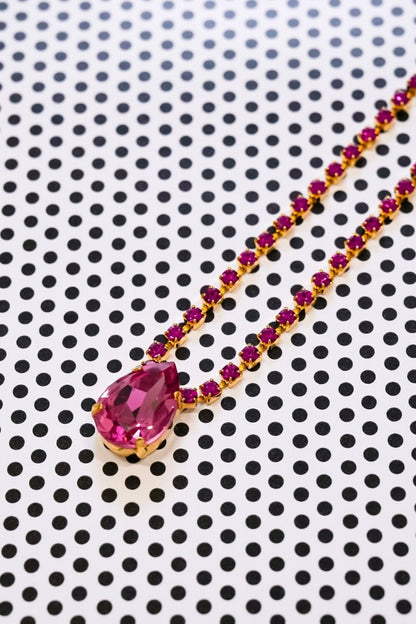 A gold necklace with a large teardrop-shaped pink gemstone pendant and a chain lined with small pink gemstones, displayed on a white background with a black polka dot pattern.