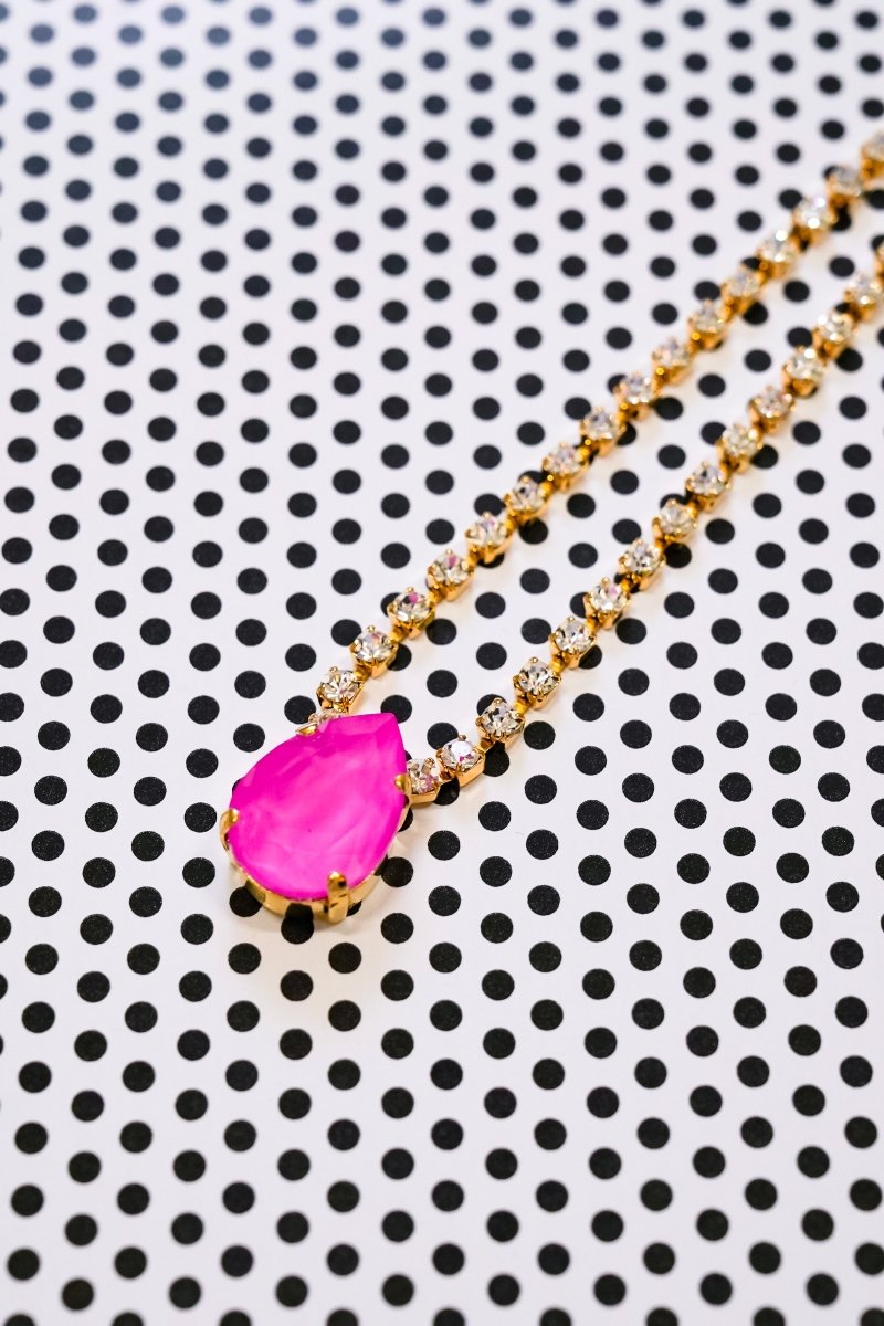 A gold chain necklace featuring a vibrant hot pink teardrop-shaped gemstone pendant, displayed diagonally on a white background with a black polka dot pattern.