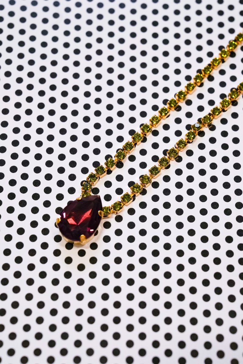 A gold chain necklace with a dark red teardrop-shaped gemstone pendant, displayed diagonally on a white background with a black polka dot pattern.