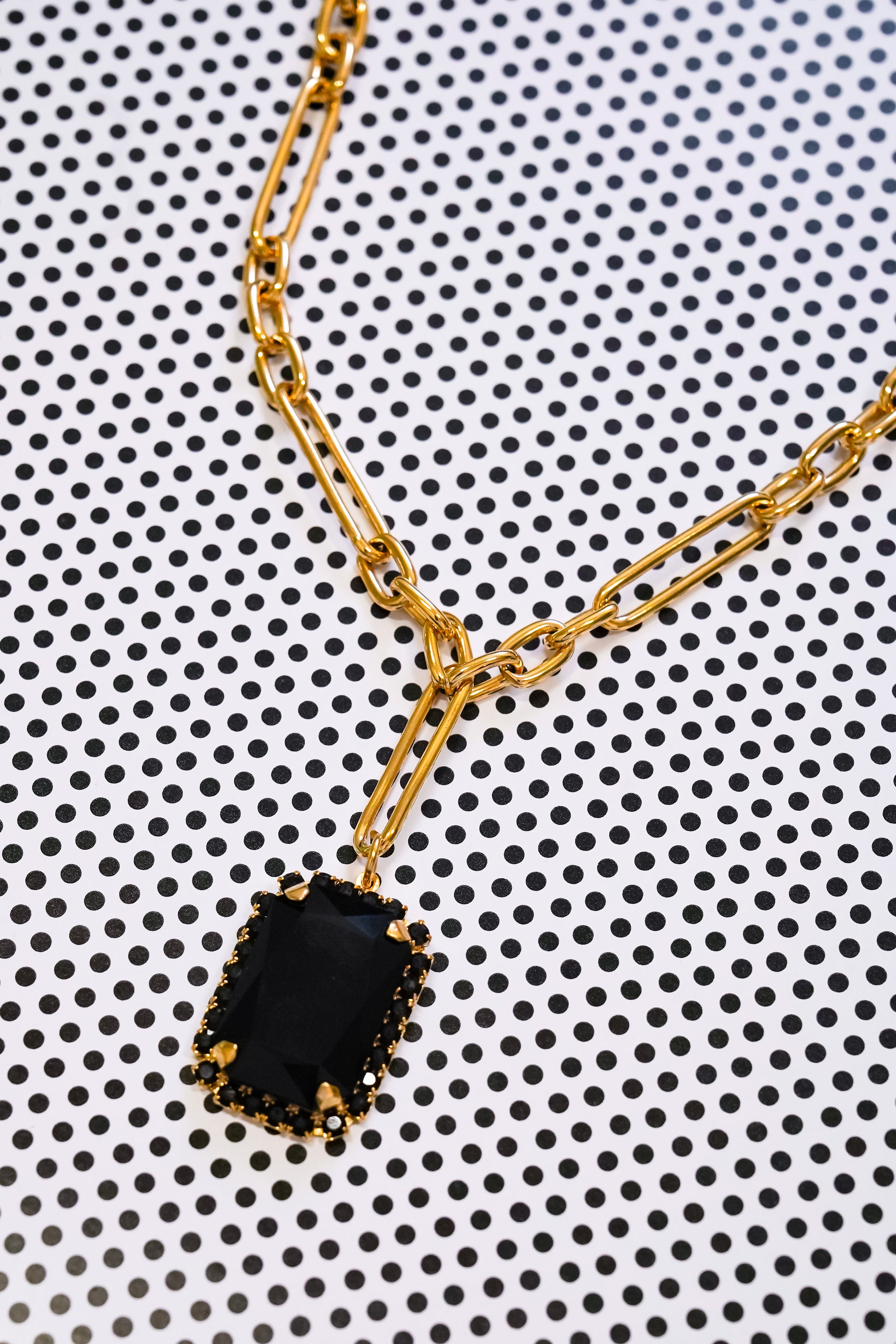 A gold chain necklace with a rectangular black pendant, photographed close-up on a white background with a black polka dot pattern.