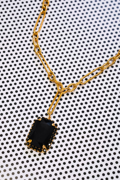 A gold chain necklace with a rectangular black pendant, photographed close-up on a white background with a black polka dot pattern.