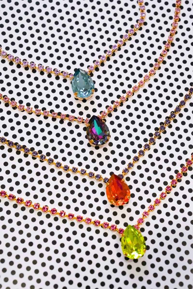 Four beaded necklaces with teardrop-shaped gemstone pendants in blue, iridescent green, orange, and lime green, arranged on a white background with a black polka dot pattern.