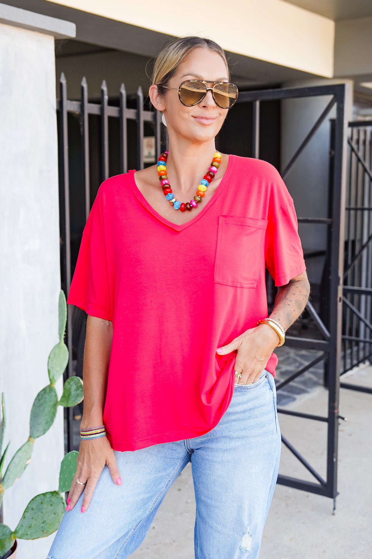 A woman wearing a bright coral pink short-sleeve V-neck top with a chest pocket, paired with light blue jeans, sunglasses, and a colorful beaded necklace, standing outdoors.