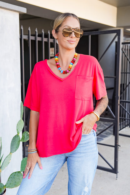 A woman wearing a bright coral pink short-sleeve V-neck top with a chest pocket, paired with light blue jeans, sunglasses, and a colorful beaded necklace, standing outdoors.