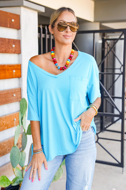 A woman wearing a light blue off-the-shoulder V-neck top with a chest pocket, paired with light-wash jeans, a colorful beaded necklace, and sunglasses, standing outdoors.