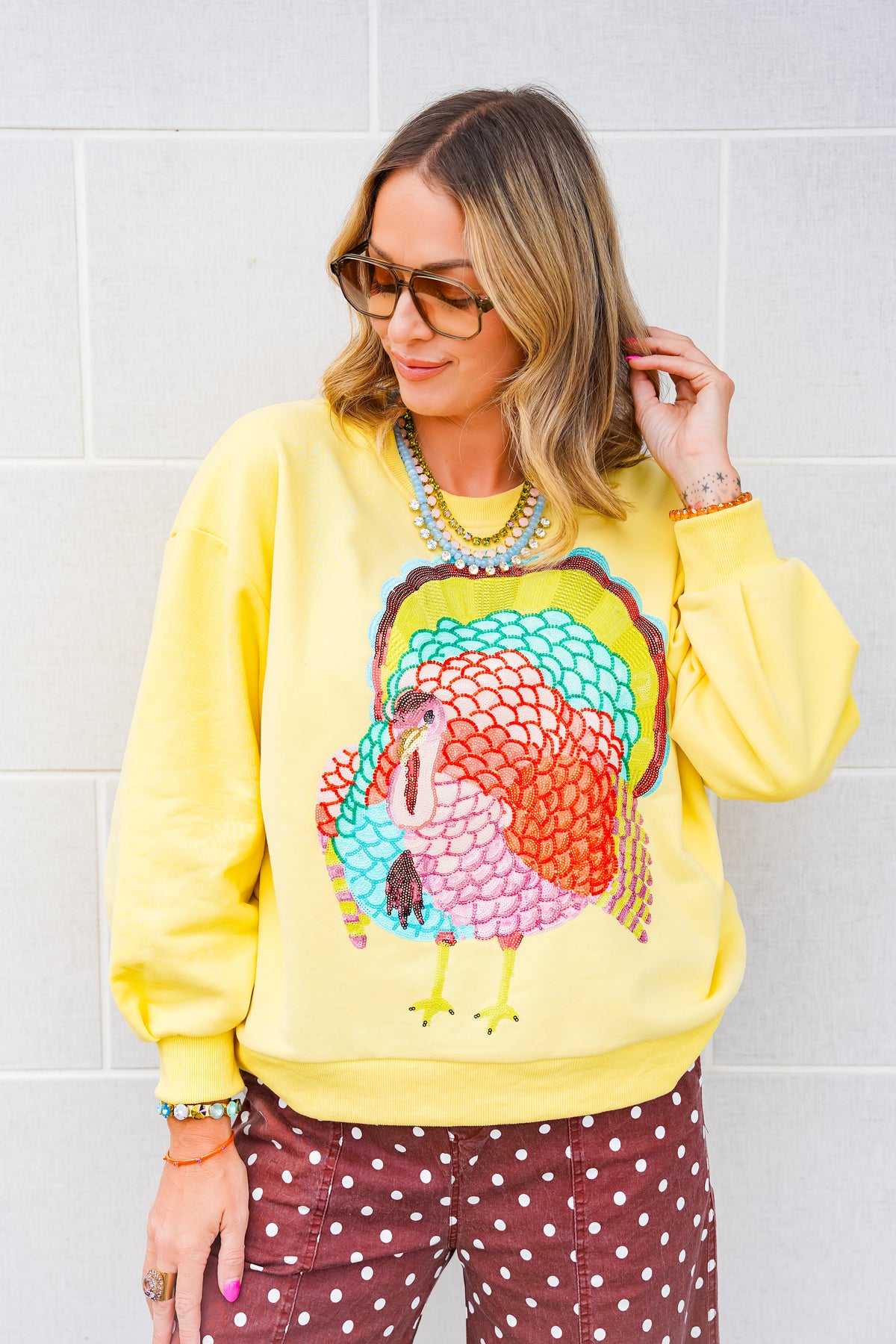 A woman wearing a bright yellow crewneck sweatshirt with a large, colorful embroidered turkey graphic on the front, paired with brown polka-dot pants, standing against a light gray brick wall.