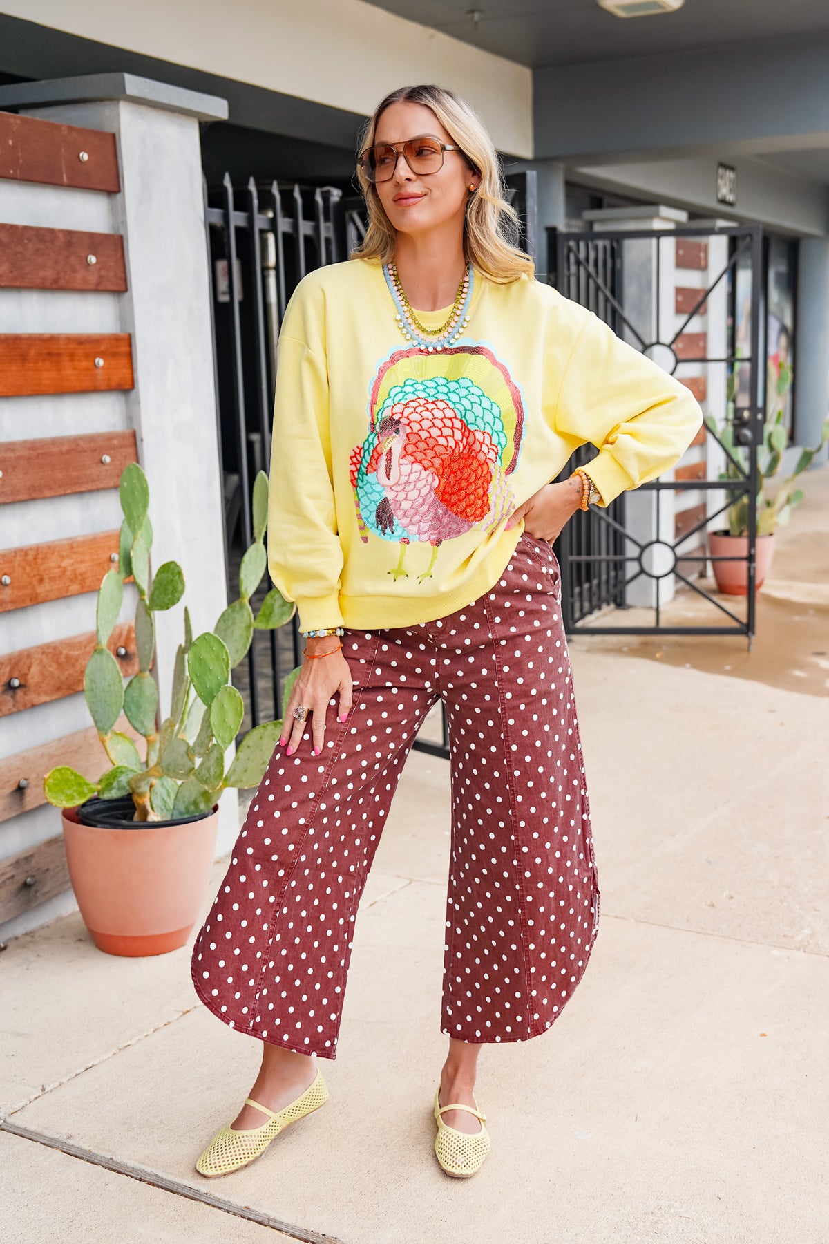 A woman modeling a yellow sweatshirt with a colorful turkey graphic, paired with brown wide-leg pants featuring white polka dots, standing on a concrete patio.