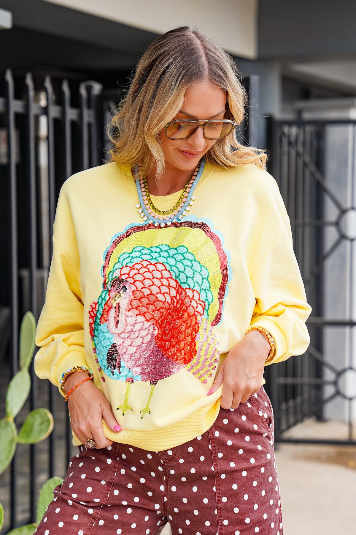 A woman wearing a bright yellow crewneck sweatshirt with a large colorful turkey graphic on the front, paired with brown polka-dot pants, standing outdoors in front of a black metal gate.