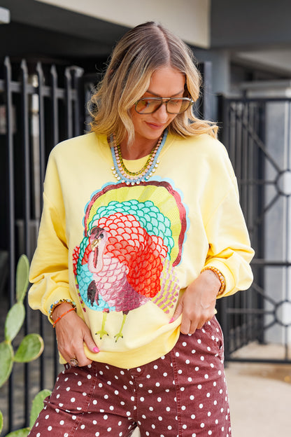 A woman wearing a bright yellow crewneck sweatshirt with a large colorful turkey graphic on the front, paired with brown polka-dot pants, standing outdoors in front of a black metal gate.