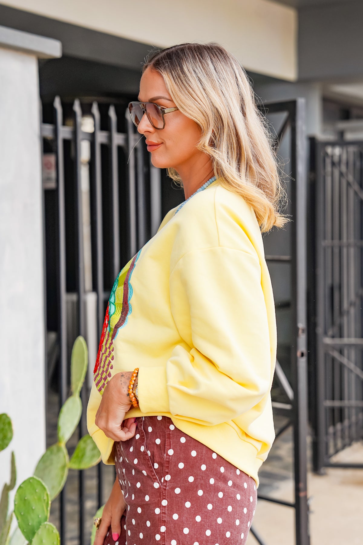 A woman wearing a pastel yellow sweatshirt with a colorful graphic on the front, paired with brown polka-dot pants, standing outdoors in front of a gate.