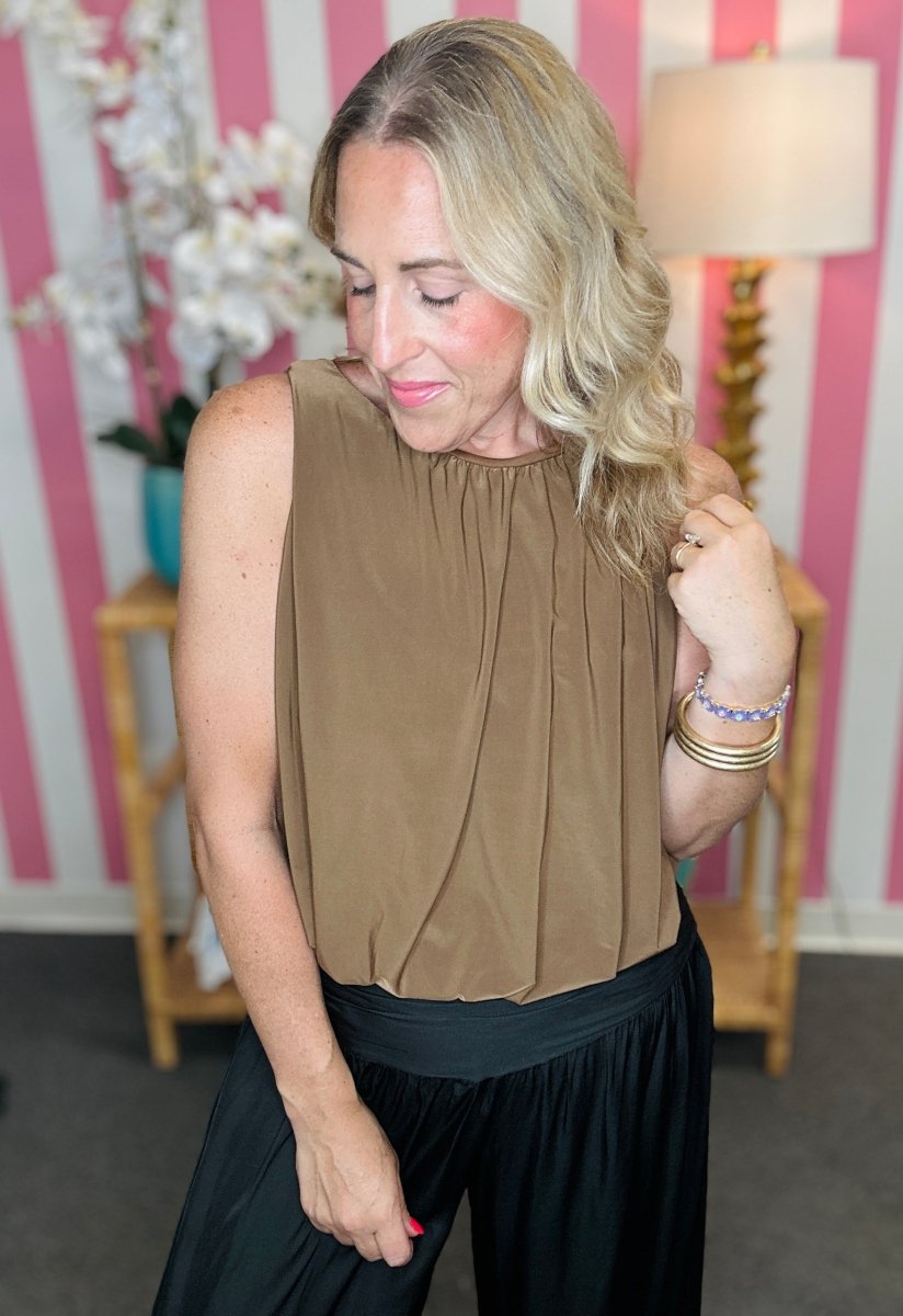 A woman wearing a sleeveless taupe-brown pleated top and black pants, standing in front of a pink and white striped wall with a lamp and side table in the background.
