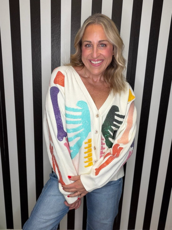 A smiling blonde woman wearing a cream-colored cardigan with colorful abstract brushstroke patterns in purple, light blue, black, yellow, and orange, paired with blue jeans, standing against a black and white vertically striped background.