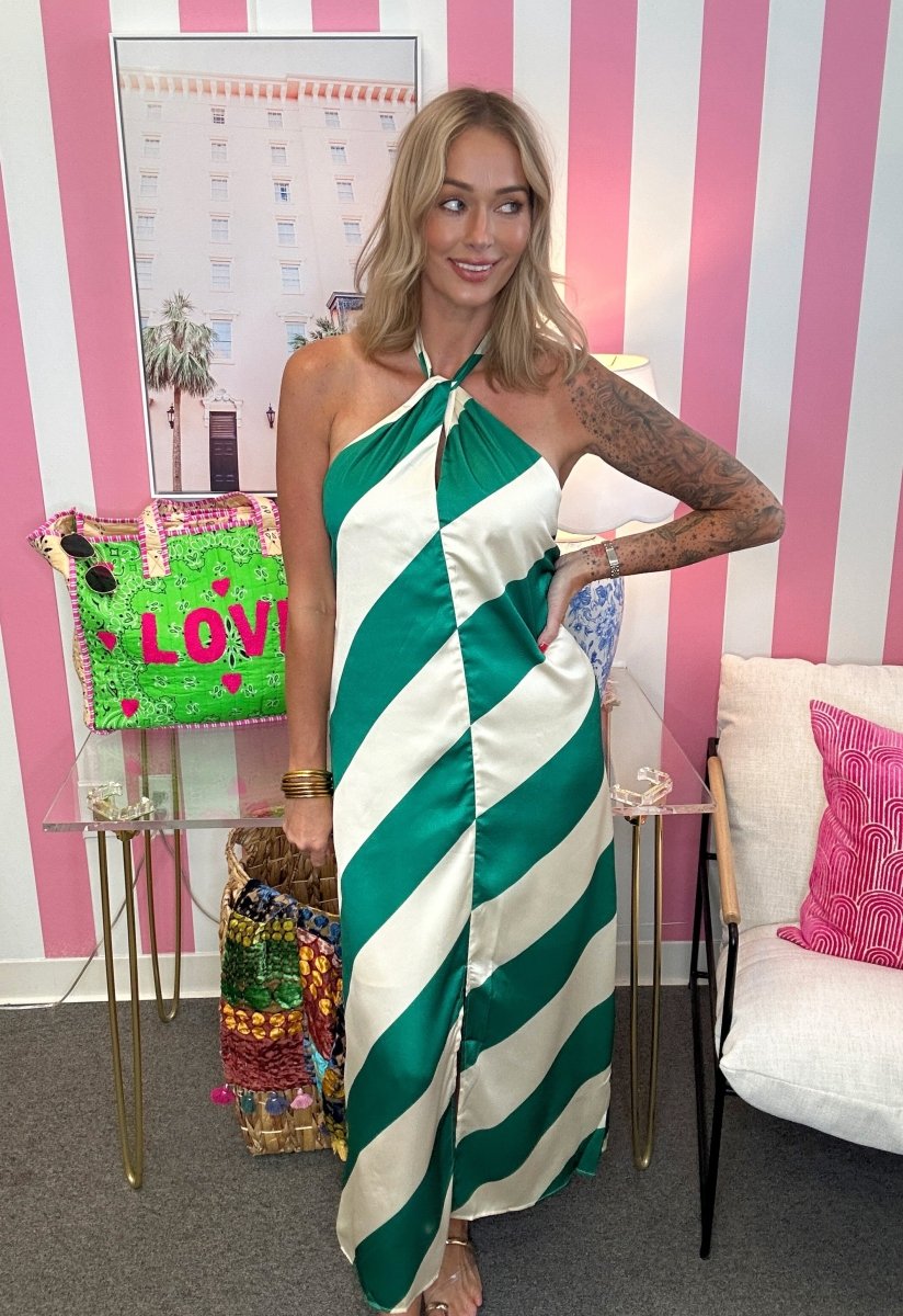 A woman wearing a green and white striped halter-neck maxi dress, posing in front of a pink and white striped wall with a glass table and a white chair.