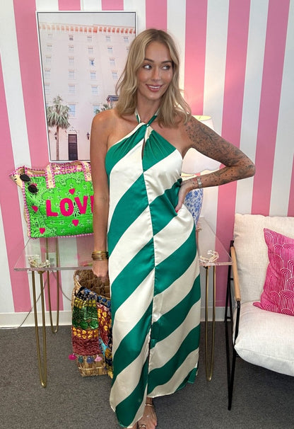 A woman wearing a green and white striped halter-neck maxi dress, posing in front of a pink and white striped wall with a glass table and a white chair.