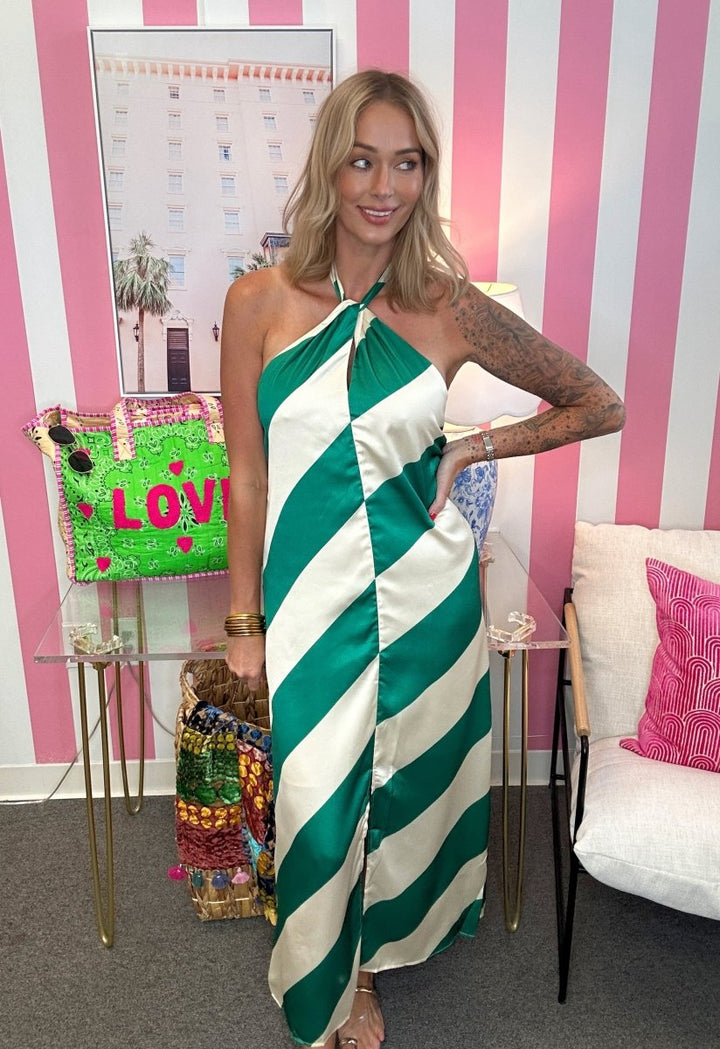 A woman wearing a green and white striped halter-neck maxi dress, posing in front of a pink and white striped wall with a glass table and a white chair.