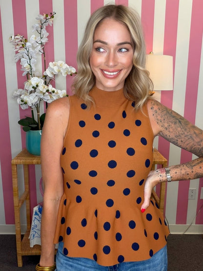A woman wearing a sleeveless mock-neck top in rust orange with navy blue polka dots, standing in front of a pink and white striped wall.