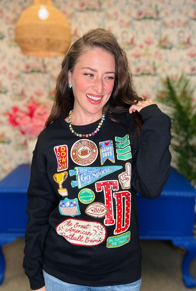 A woman smiling and posing in a black crewneck sweatshirt decorated with various colorful football-themed patches and text, set against a floral wallpaper background with a blue table.