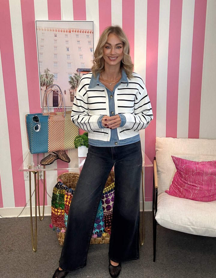 A woman stands in a pink and white striped room wearing a white cropped cardigan with black horizontal stripes and a light blue collar and button placket, paired with dark wide-leg jeans.