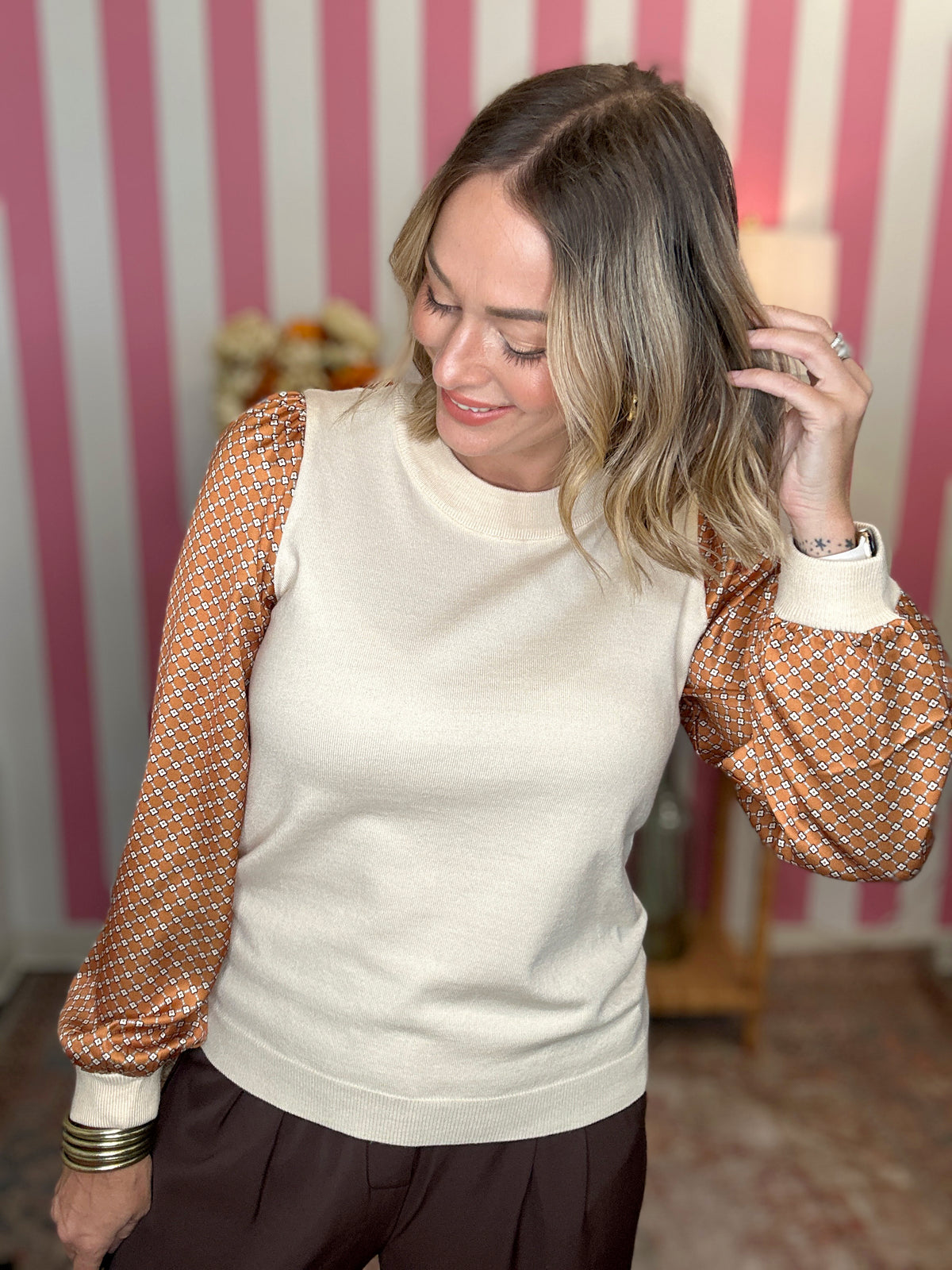 A woman wearing a cream knit sweater vest with patterned balloon sleeves, posed in front of a pink and white striped wall.
