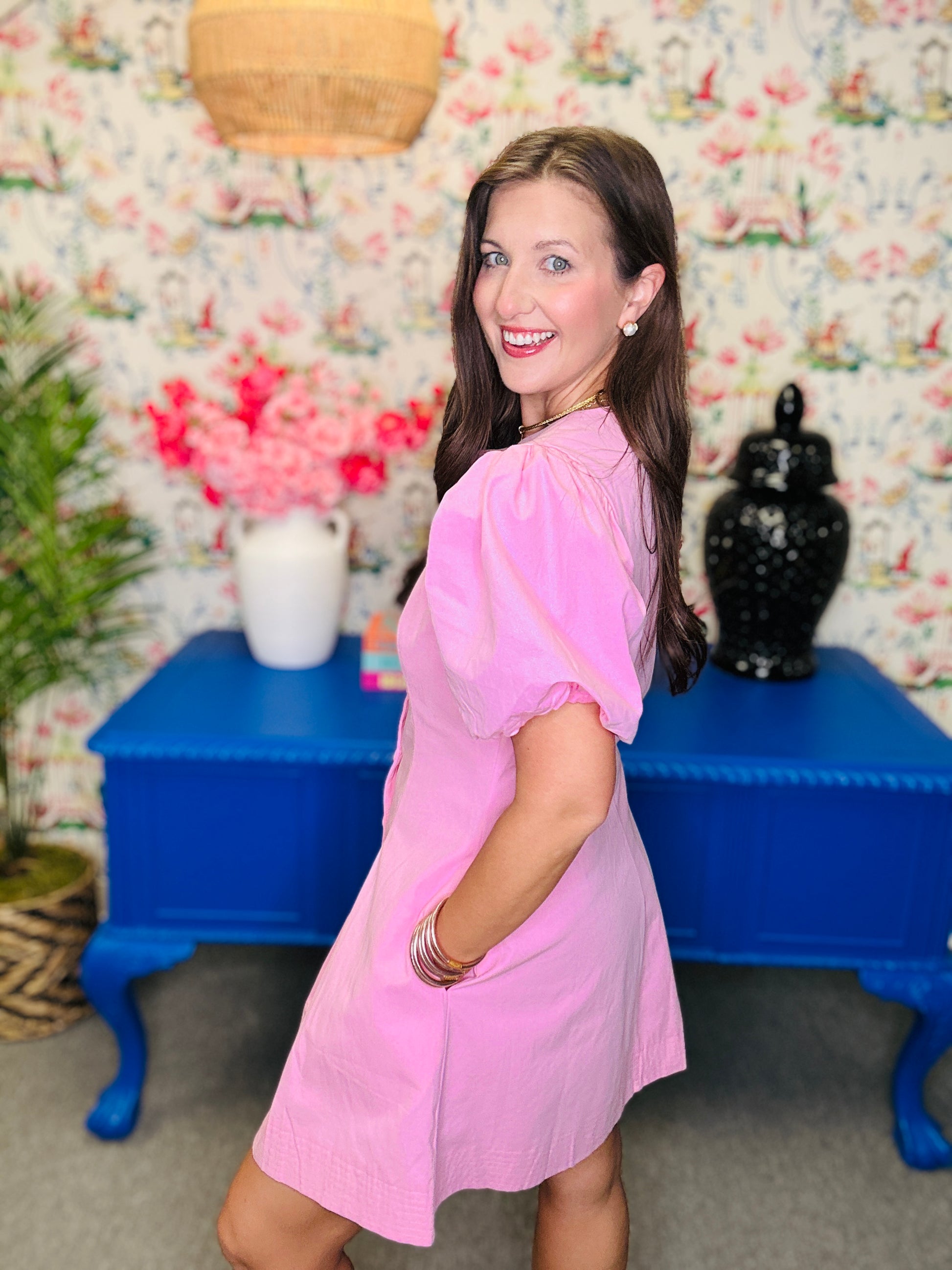 Woman in a pink dress standing in front of a colorful floral wall with a blue table and decorative items.
