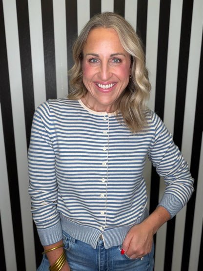 A smiling blonde woman wearing a blue and cream striped button-front cardigan over a light top and blue jeans, standing in front of a black and white vertically striped wall.