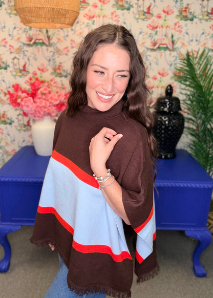 A woman wearing a brown, light blue, and red striped shawl poncho in a decorated room with floral wallpaper and a blue side table.