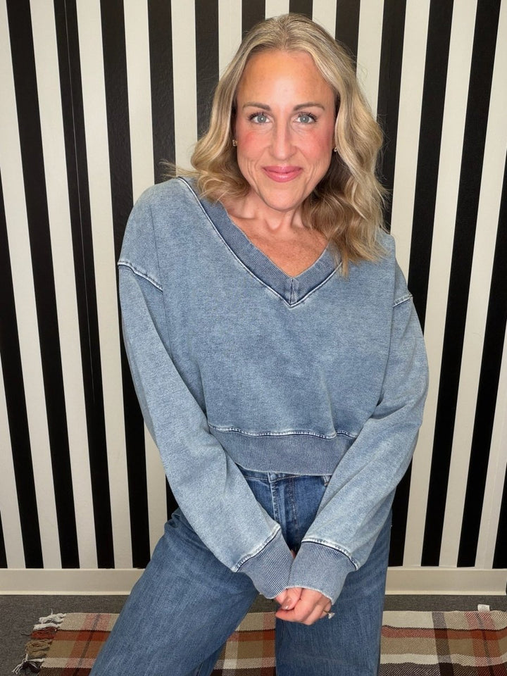 A woman with blonde hair wearing a faded light blue cropped V-neck long-sleeve top and blue jeans, posing in front of a black and white striped wall.