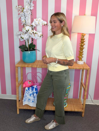 A woman modeling a pale yellow and white striped mock neck top with olive green cuffed pants and metallic silver sneakers, standing in front of a pink and white striped wall next to a bamboo console table with a lamp and a potted orchid.