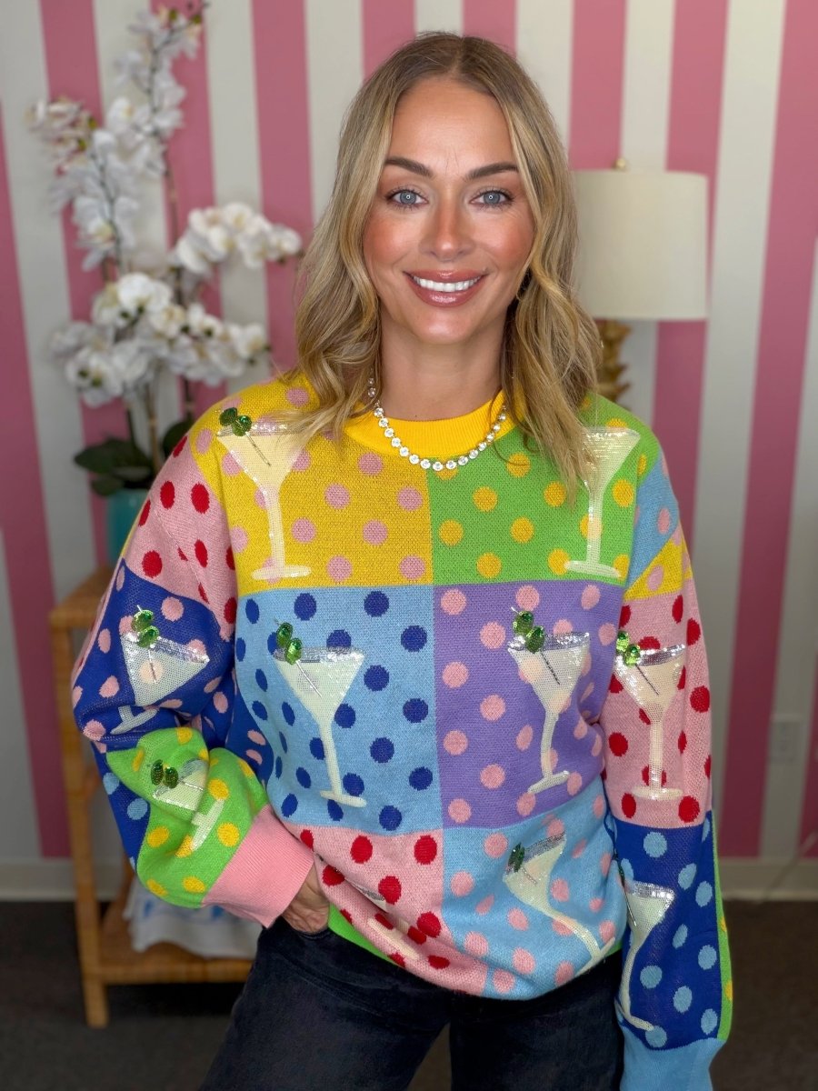 A woman wearing a vibrant, multicolored knit sweater with a patchwork design featuring cocktail glasses and polka dots, posed in front of a pink and white striped wall.