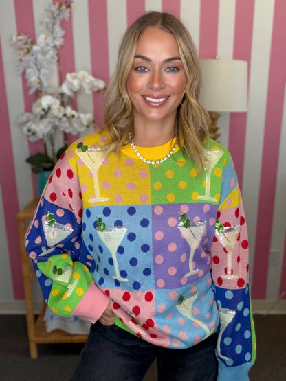 A woman wearing a vibrant, multicolored knit sweater with a patchwork design featuring cocktail glasses and polka dots, posed in front of a pink and white striped wall.