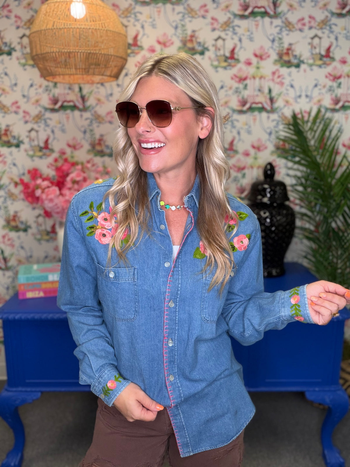 A woman wearing a light blue denim button-up shirt with pink floral embroidery on the chest and cuffs, styled with brown pants and sunglasses, standing in front of a floral wallpaper background.