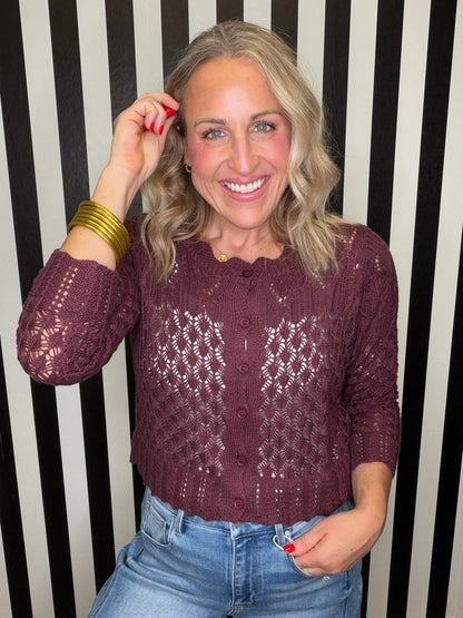 A smiling blonde woman wearing a maroon open-knit long-sleeve top and blue jeans, standing against a black and white vertically striped background.