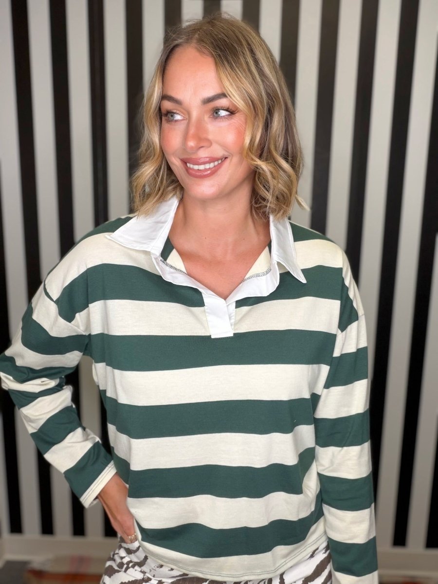A woman wearing a green and off-white horizontally striped long-sleeve rugby shirt, standing in front of a black and white striped background.