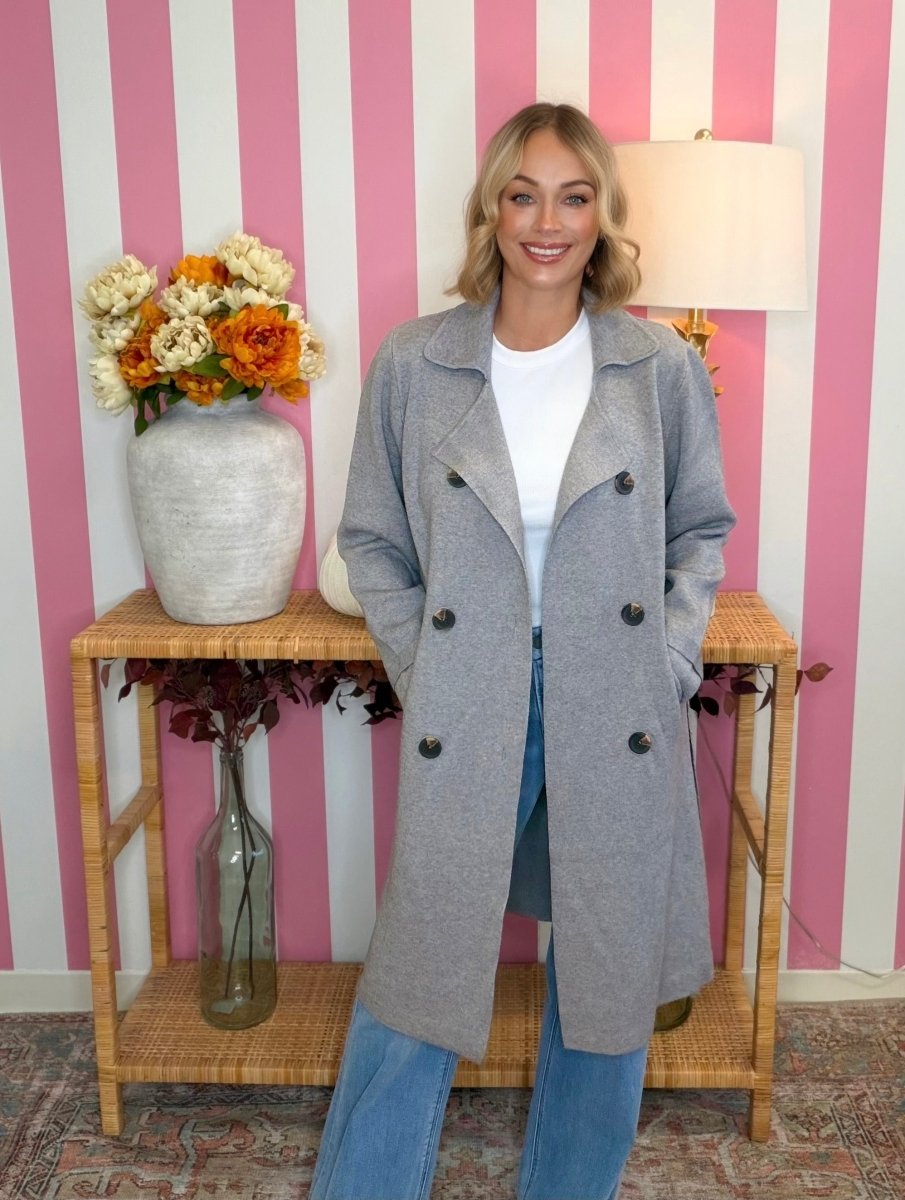 A woman wearing a light gray double-breasted coat over a white top and blue jeans, standing in front of a pink and white striped wall with a wicker console table and a vase of flowers.