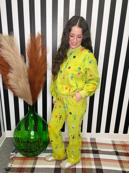 A woman modeling a bright yellow-green matching sweatshirt and sweatpants set with a colorful paint splatter print, standing in front of a black and white striped wall next to a green glass vase with pampas grass.