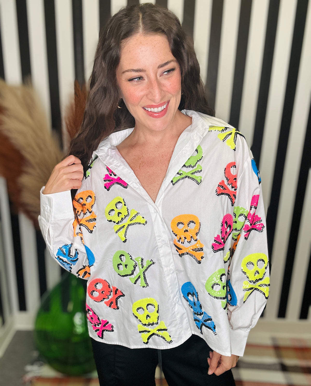 A woman smiling while wearing a white button-up shirt covered in colorful skull and crossbones motifs, paired with black pants, standing in front of a black and white striped background.
