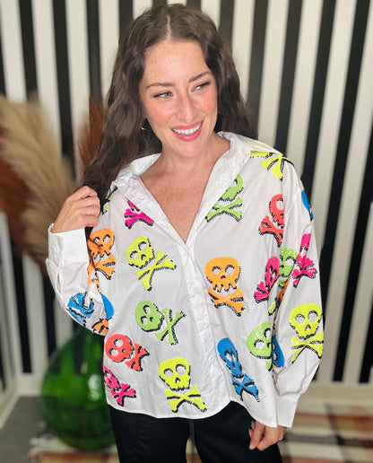 A woman smiling while wearing a white button-up shirt covered in colorful skull and crossbones motifs, paired with black pants, standing in front of a black and white striped background.
