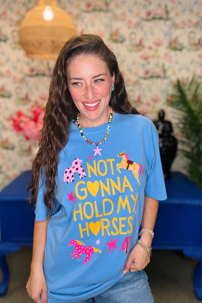 A woman wearing a light blue graphic t-shirt with the text 'NOT GONNA HOLD MY HORSES' in yellow, decorated with pink and yellow horses. She is smiling and posing in front of a floral wallpaper background.