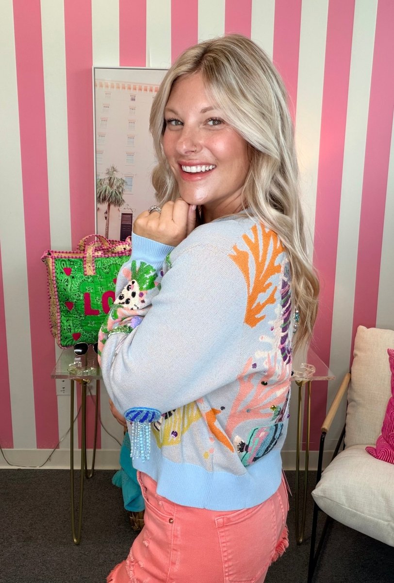 Woman in a colorful sweater holding a green bag with a character design, standing against a striped pink and white wall.