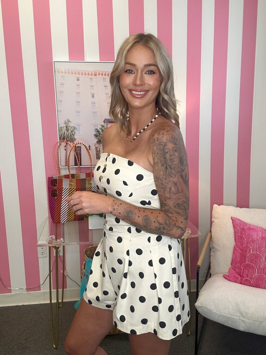 Woman in a polka dot dress holding a colorful handbag against a pink and white striped wall.