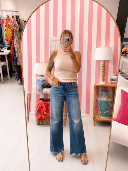 Person wearing a white tank top and blue jeans standing in front of a mirror with a pink and white striped wall.