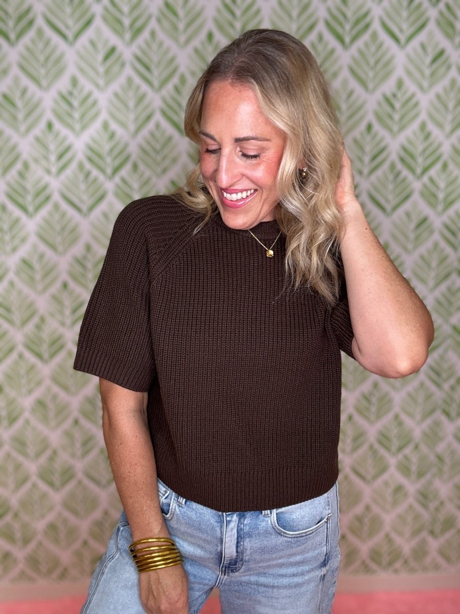 A woman with blonde hair wearing a brown short-sleeve ribbed knit top and light blue jeans, standing in front of a green and white patterned wallpaper background.