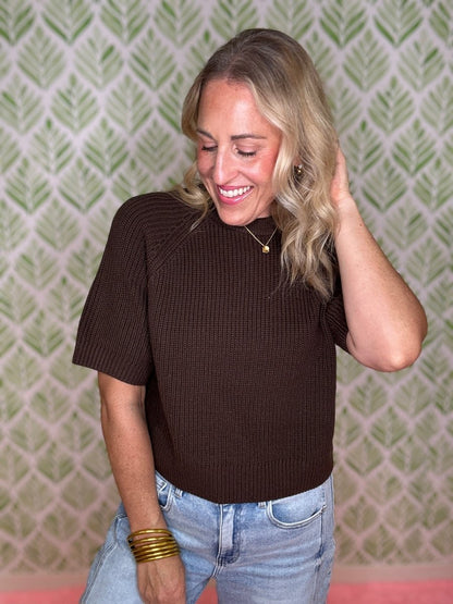 A woman with blonde hair wearing a brown short-sleeve ribbed knit top and light blue jeans, standing in front of a green and white patterned wallpaper background.