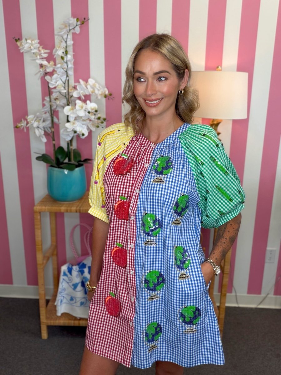 A woman modeling a short puff-sleeve dress with a vertical multicolor gingham split: red/white, blue/white, and green/white panels with fruit and globe appliques, posed in front of pink-and-white striped wallpaper.