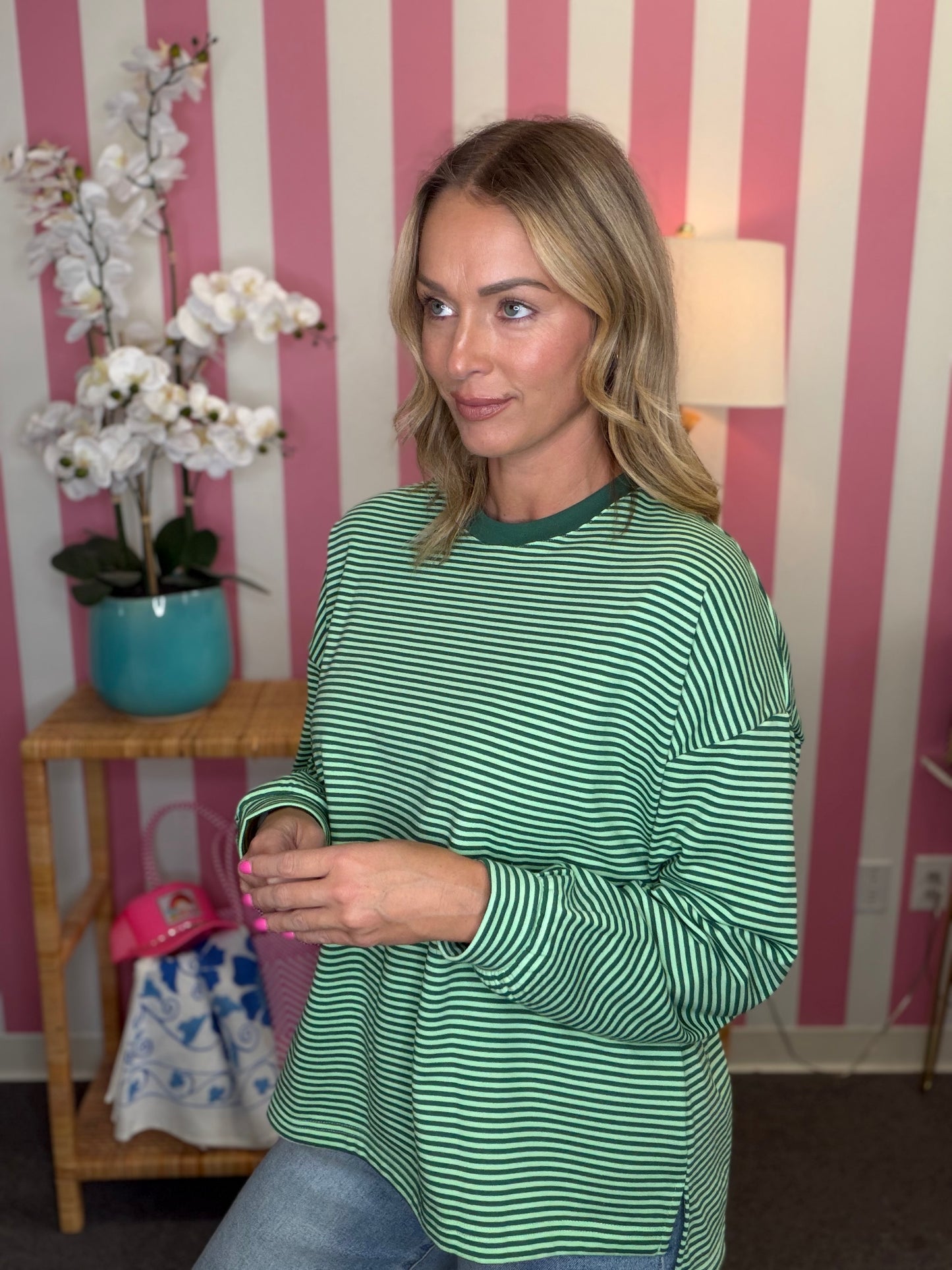 A woman wearing a green and white striped long-sleeve top, standing in front of a pink and white striped wall with a small wooden side table and white flowers in the background.