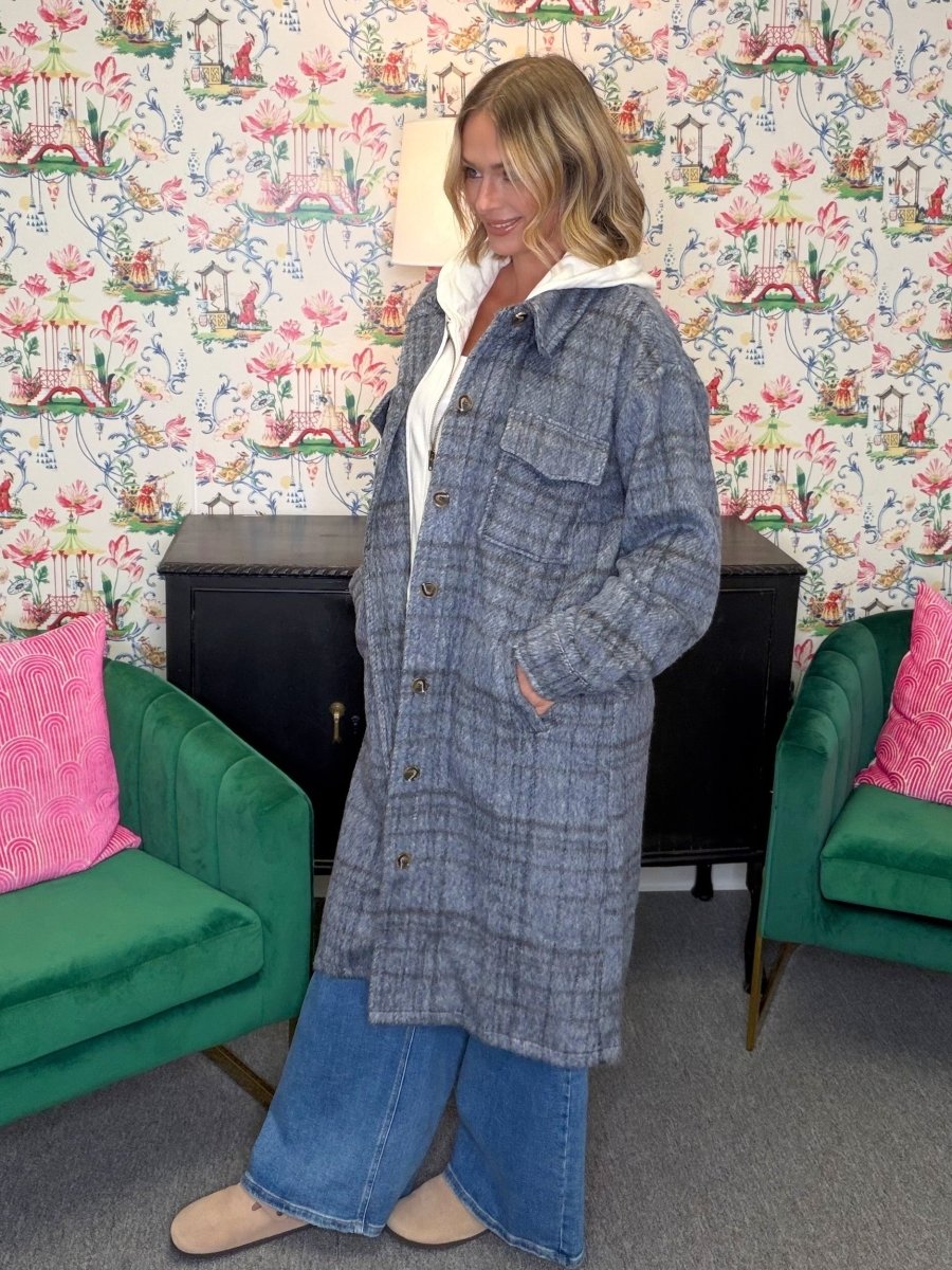 A woman wearing a long gray plaid coat over a white hoodie and blue jeans, standing in a room with floral wallpaper, green armchairs, and a black cabinet.